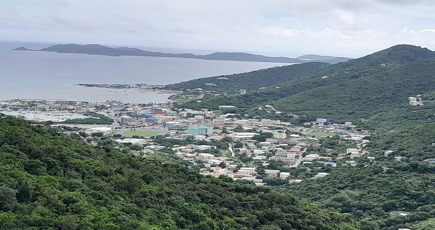 Rich History of the Virgin Islands. An elevated view looking down a steep, green, densely-forested valley toward a coastal city of Road Town, Tortola clustered around a harbor, with the blue ocean and distant islands in the background.