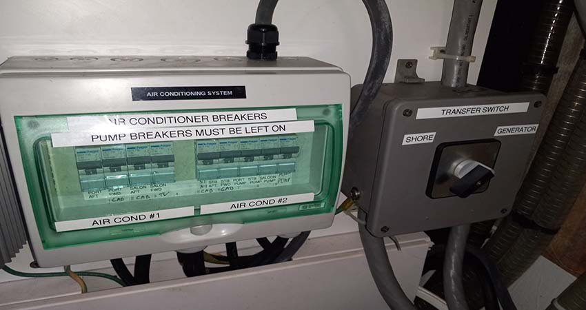 Essential Yacht Equipment. A close-up of a yacht's electrical panel, showing an air conditioning breaker box with labeled switches and a transfer switch labeled SHORE and GENERATOR. class=