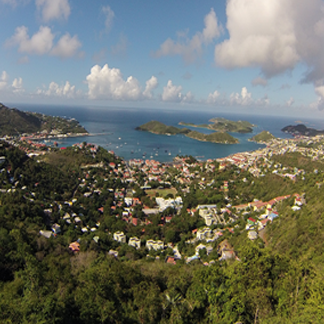 A wide, elevated panoramic view of a densely built-up coastal city of Charlotte Amalie, St. Thomas nestled around a curving harbor filled with boats. The bright blue ocean, distant islands, and a partly cloudy sky complete the scene.