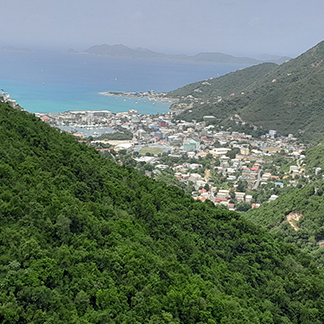 An elevated view looking down a steep, green, densely-forested valley toward the coastal city of Road Town, Tortola clustered around a harbor, with the blue ocean and distant islands in the background.